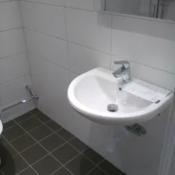 Biała umywalka z chromowaną baterią i odpływem, zamontowana na ścianie z białych płytek, z instrukcją obsługi obok baterii, szare płytki podłogowe, widok na kabinę prysznicową w lustrze.