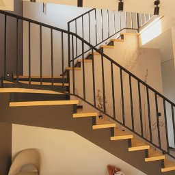 Steel Masters - Nowoczesne schody z drewnianymi stopniami i czarną metalową balustradą, widok z dołu, w tle osoba sznuruje buty przy białych drzwiach.