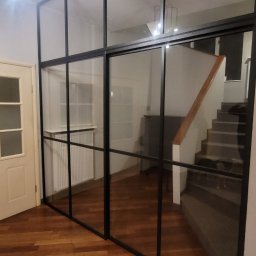 Steel Masters - Przeszklona ścianka działowa z czarnymi profilami oddzielająca korytarz ze schodami od pokoju z parkietem i grzejnikiem.