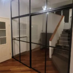 Przeszklona ścianka działowa z czarnymi profilami oddzielająca korytarz ze schodami od pokoju z parkietem i grzejnikiem.