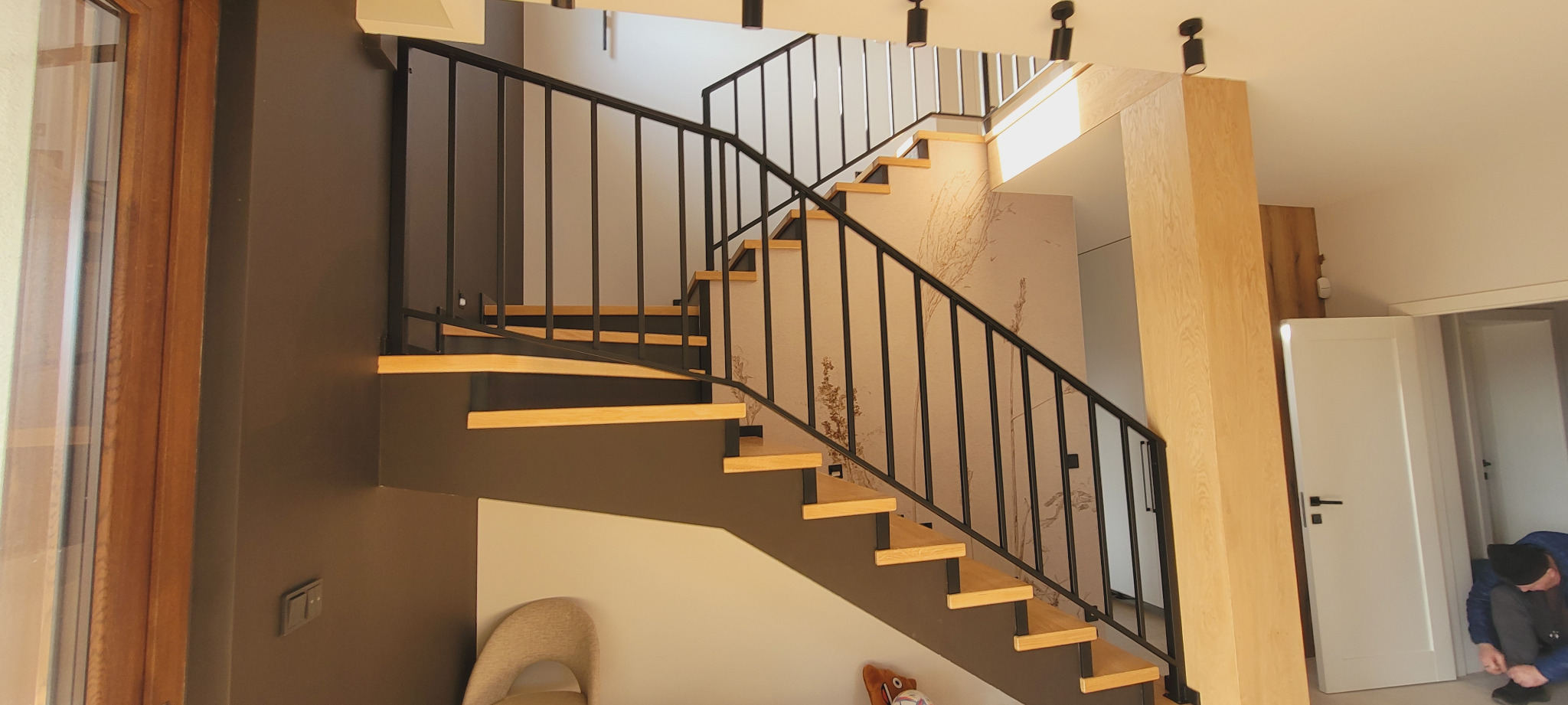 Nowoczesne schody z drewnianymi stopniami i czarną metalową balustradą, widok z dołu, w tle osoba sznuruje buty przy białych drzwiach.