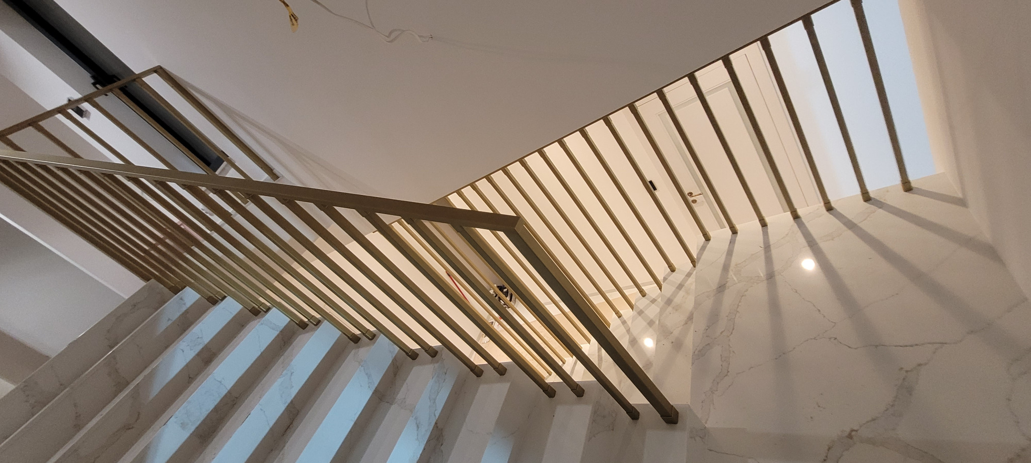 Nowoczesne schody z marmurowymi stopniami i złotą, minimalistyczną balustradą z pionowych prętów, widok z dołu w górę, oświetlenie punktowe odbijające się w polerowanym kamieniu.