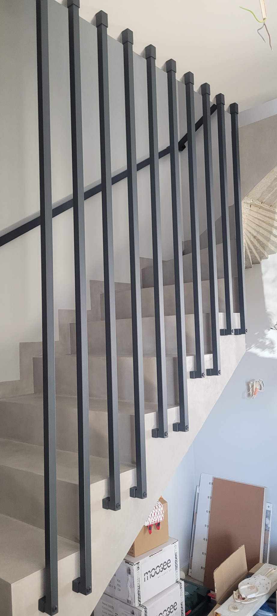 Nowoczesna klatka schodowa z minimalistyczną, ciemnoszarą balustradą z pionowych słupków o kwadratowym przekroju, widoczne surowe wykończenie ścian i schodów, w tle elementy budowlane i kartony.