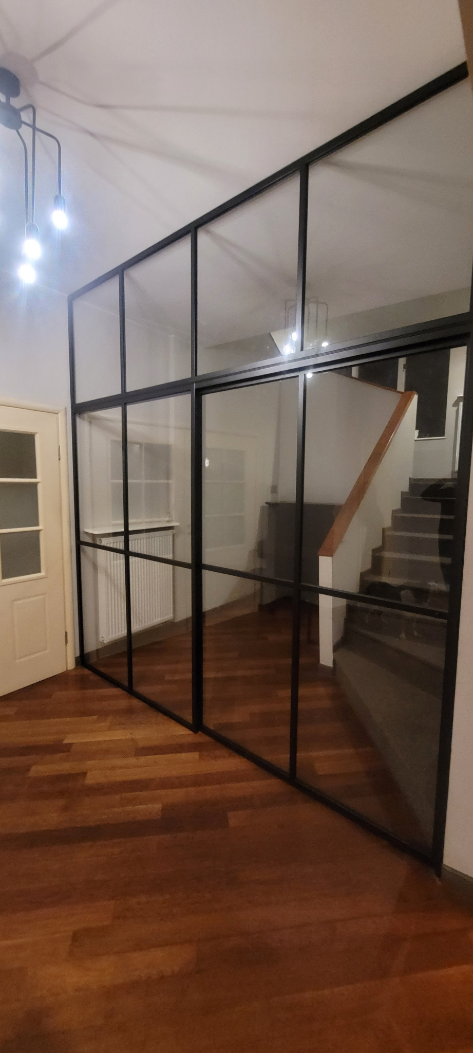 Przeszklona ścianka działowa z czarnymi profilami oddzielająca korytarz ze schodami od pokoju z parkietem i grzejnikiem.