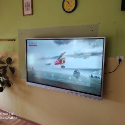 Em-Group Multimedia Sp. z o. o. - Interaktywna tablica myBoard z włączonym ekranem głównym, zawieszona na żółtej ścianie, obok doniczki z kwiatem, widoczny zegar i gniazdko elektryczne.