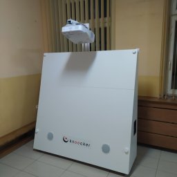 Em-Group Multimedia Sp. z o. o. - Biała obudowa interaktywnego urządzenia prezentacyjnego z logo 'knooker' i zamontowanym projektorem Epson na wysięgniku, ustawiona w pomieszczeniu z żaluzjami pionowymi.
