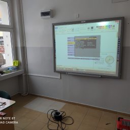 Em-Group Multimedia Sp. z o. o. - Interaktywna tablica w klasie wyświetlająca ekran programu graficznego z menu ustawień obrazu, przewód zasilający na podłodze, widok sali lekcyjnej z oknem w tle.