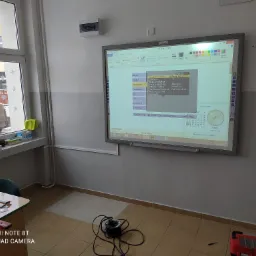 Interaktywna tablica w klasie wyświetlająca ekran programu graficznego z menu ustawień obrazu, przewód zasilający na podłodze, widok sali lekcyjnej z oknem w tle.