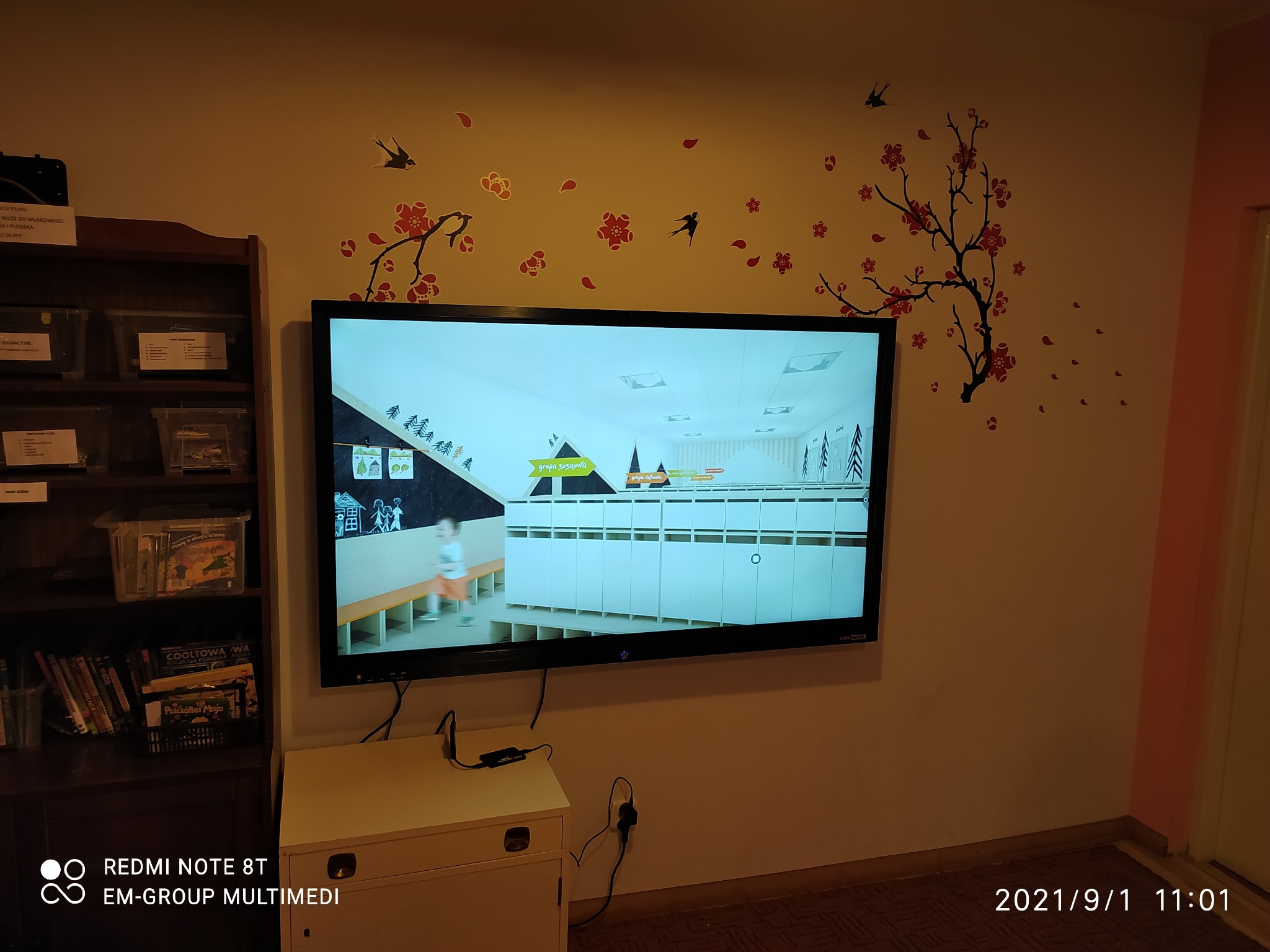 Telewizor LCD zawieszony na ścianie z naklejkami kwiatów i ptaków, wyświetlający wnętrze szatni szkolnej z szafkami i biegnącym dzieckiem.