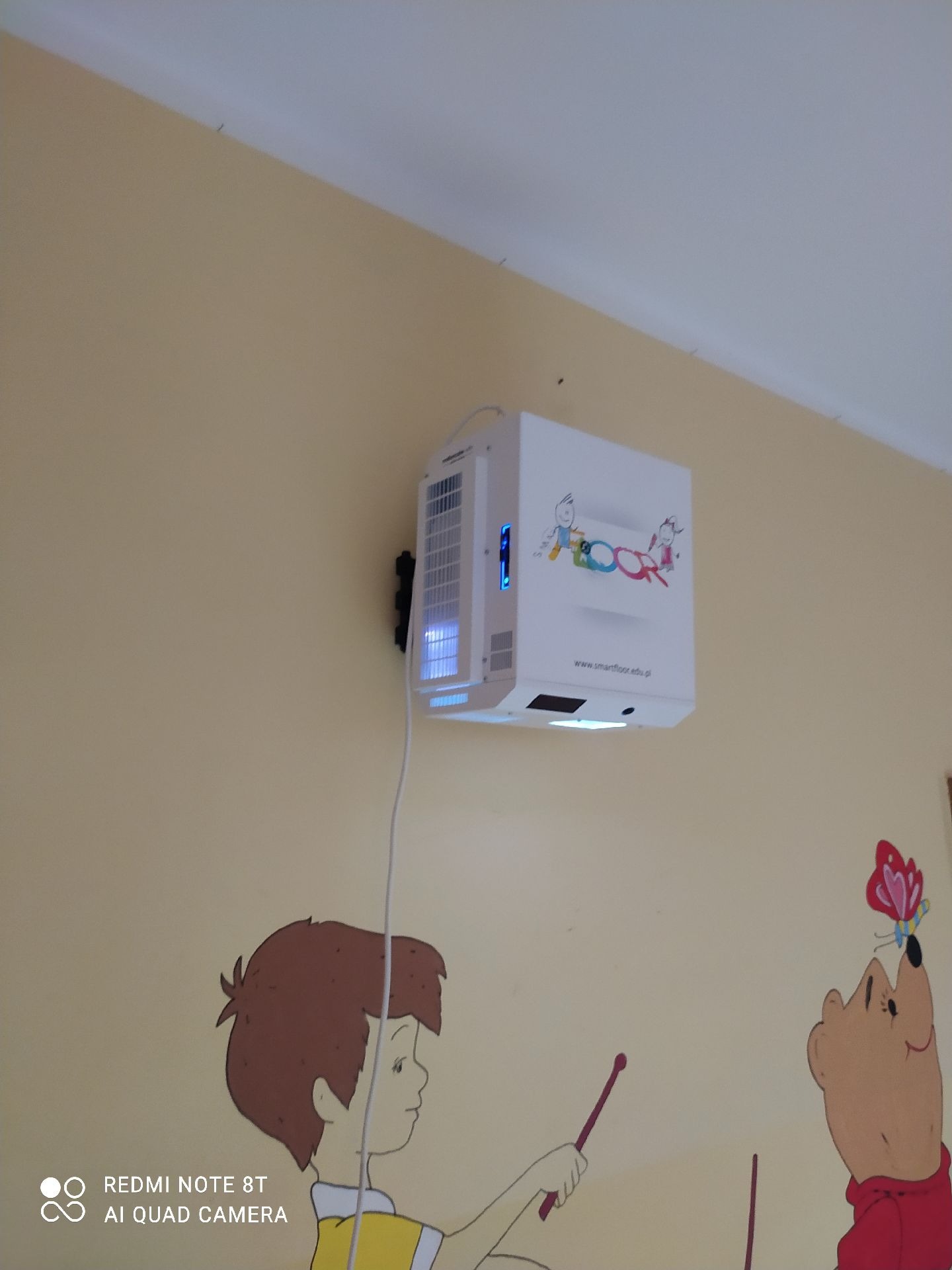 Biały projektor interaktywny zamontowany na ścianie pomalowanej w dziecięce rysunki postaci z bajek, widoczne logo i adres strony internetowej producenta.