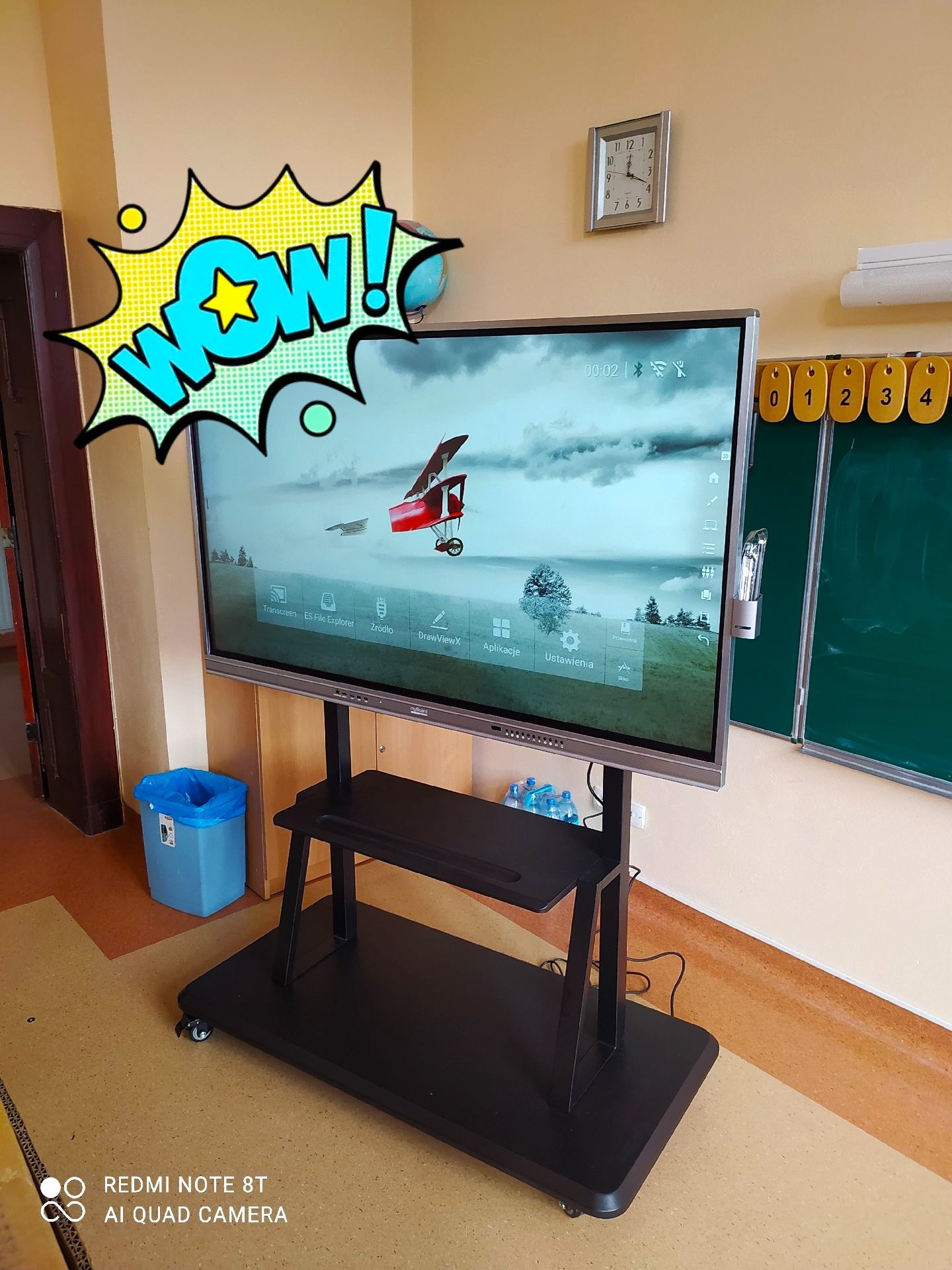 Interaktywna tablica multimedialna na mobilnym stojaku w klasie, z wyświetlanym pulpitem i ikoną 'WOW!', obok tradycyjna tablica, zegar i liczydło.