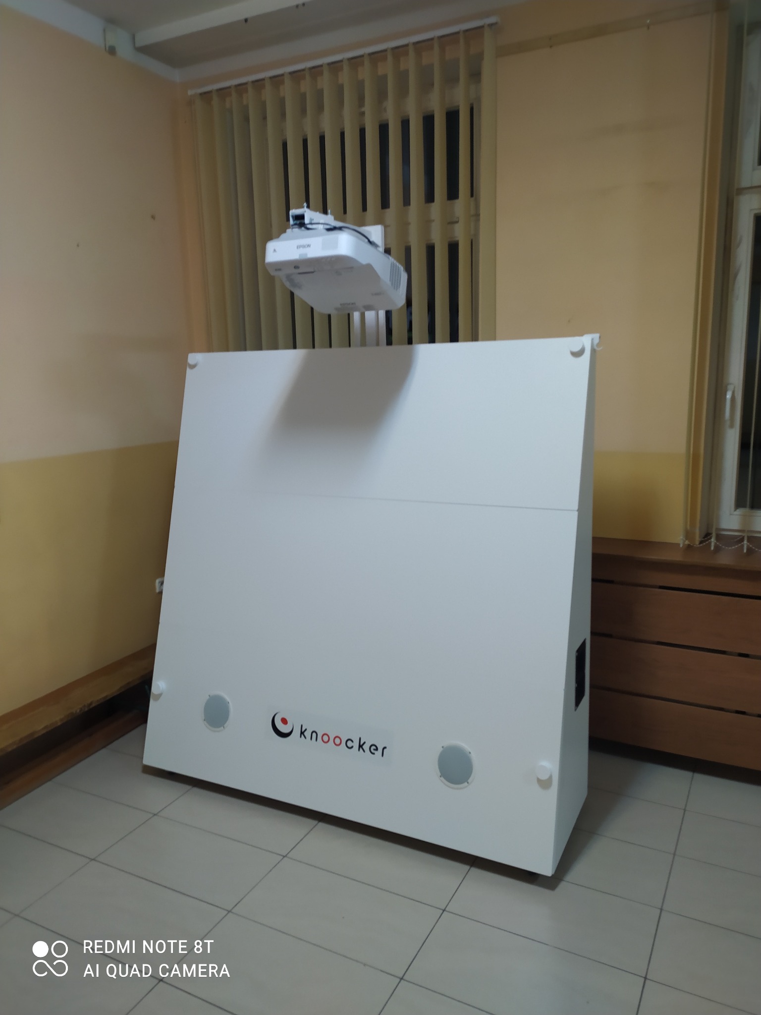Biała obudowa interaktywnego urządzenia prezentacyjnego z logo 'knooker' i zamontowanym projektorem Epson na wysięgniku, ustawiona w pomieszczeniu z żaluzjami pionowymi.