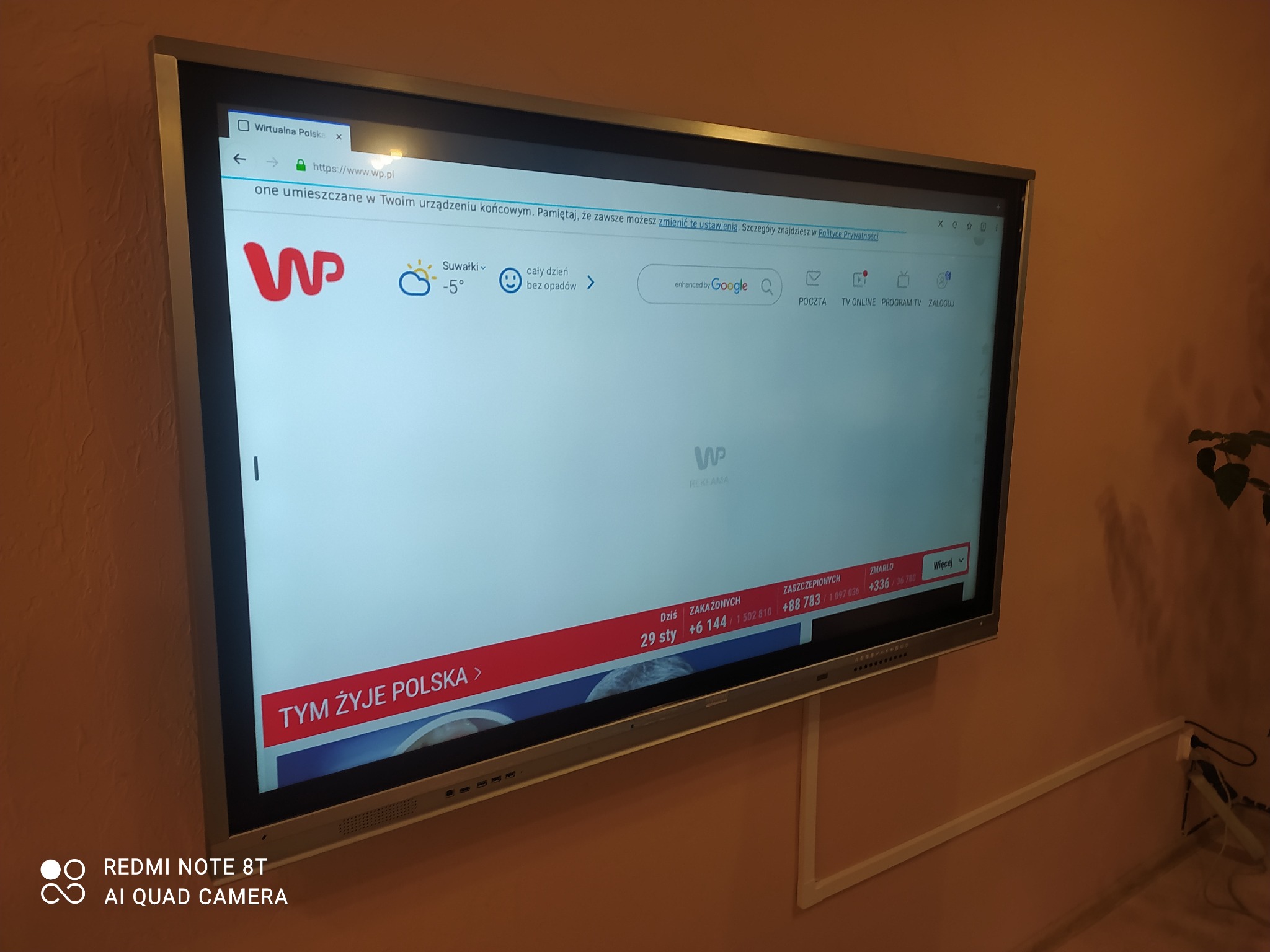 Duży ekran telewizora LCD zawieszony na ścianie w biurze, wyświetlający stronę internetową z wiadomościami i informacjami o pogodzie.