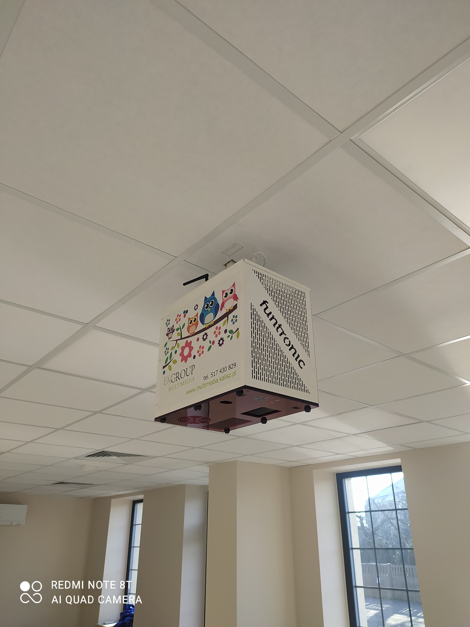 Projektor multimedialny marki Funtronic zamontowany pod sufitem kasetonowym w biurze, widoczne logo firmy EMGROUP MULTIMEDIA z motywem sów i kwiatów.