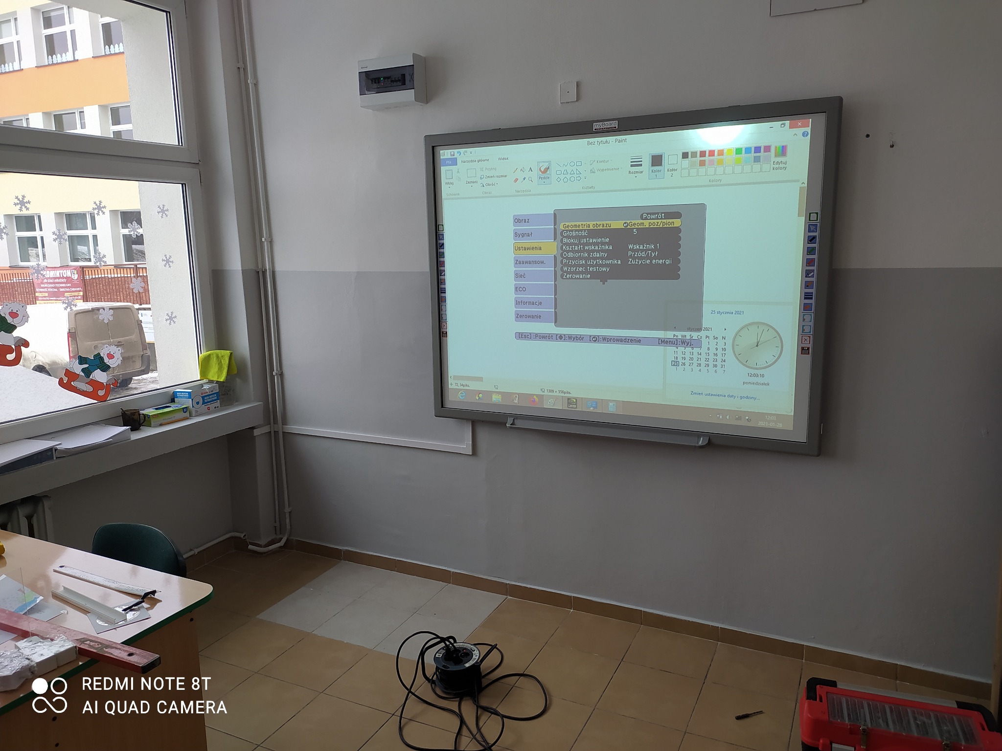 Interaktywna tablica w klasie wyświetlająca ekran programu graficznego z menu ustawień obrazu, przewód zasilający na podłodze, widok sali lekcyjnej z oknem w tle.