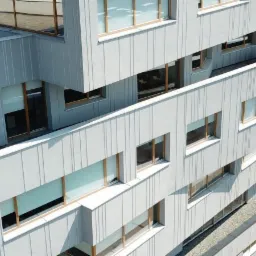 Nowoczesny budynek biurowy z szarą elewacją panelową i drewnianymi ramami okiennymi, widok z góry na fragment parkingu i otoczenie.