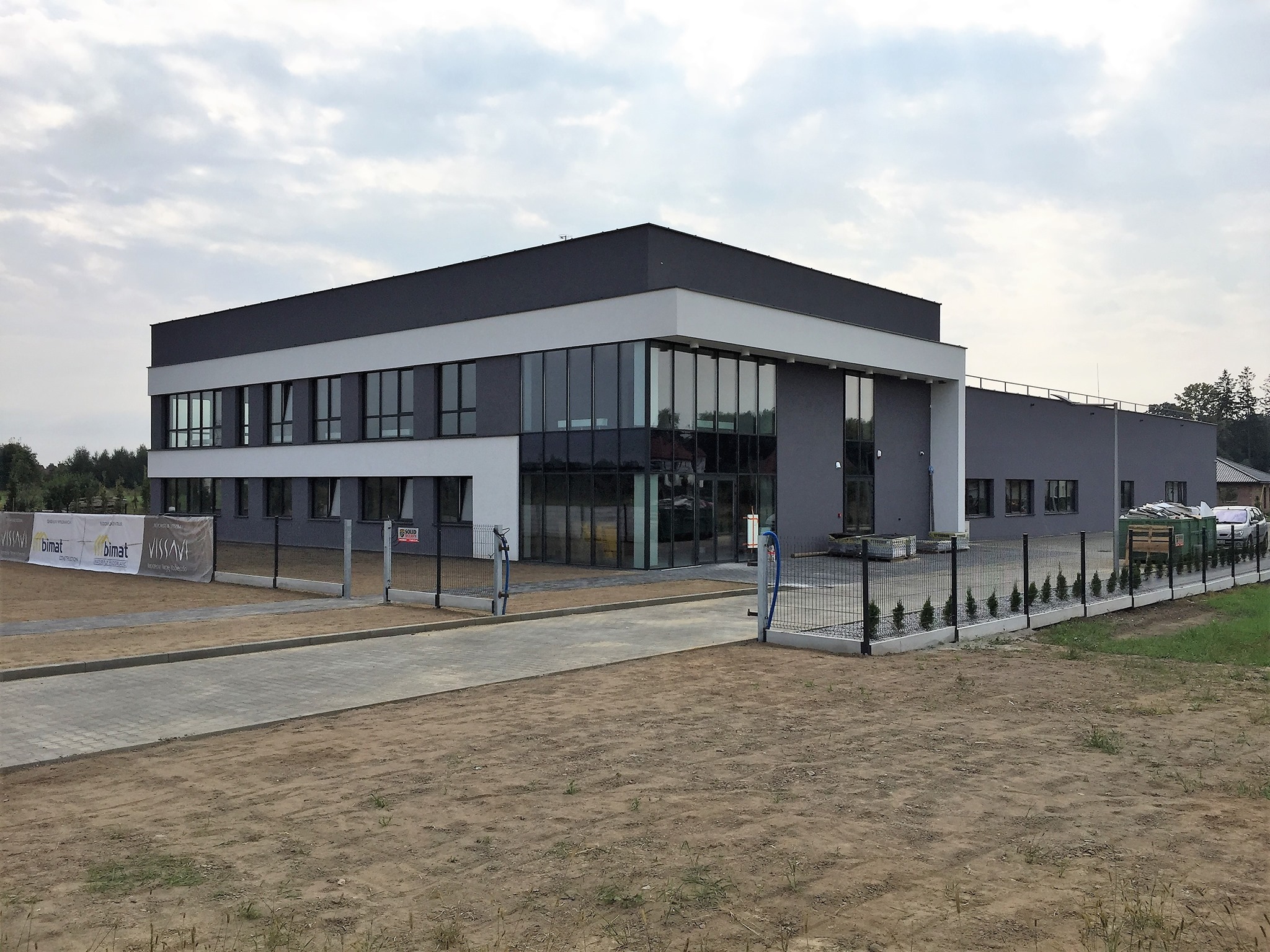 Nowoczesny budynek biurowy z szarą elewacją, dużymi oknami i płaskim dachem, otoczony trawnikiem i ogrodzeniem panelowym, widoczne banery reklamowe.