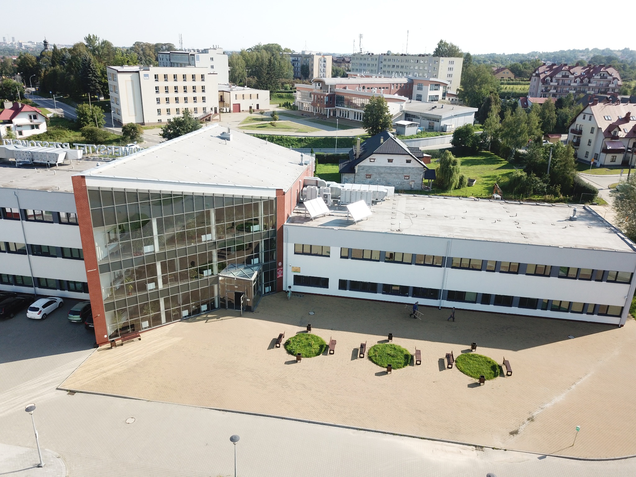 Widok z lotu ptaka na budynek biurowy z przeszkloną fasadą i elementami z czerwonej cegły, otoczony placem z zielonymi wyspami i ławkami, na tle zabudowy miejskiej w słoneczny dzień.