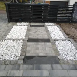 Ułożona ścieżka z szarych kostek brukowych, z obrzeżami z jasnych kamieni i białych otoczaków, prowadząca do czarnej bramy wjazdowej z automatyką.