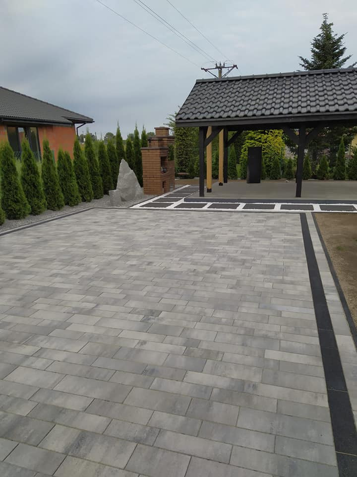 Ułożona szara kostka brukowa na podjeździe z czarnym obramowaniem, w tle altana z ciemną dachówką, ceglany grill i rząd tuj.