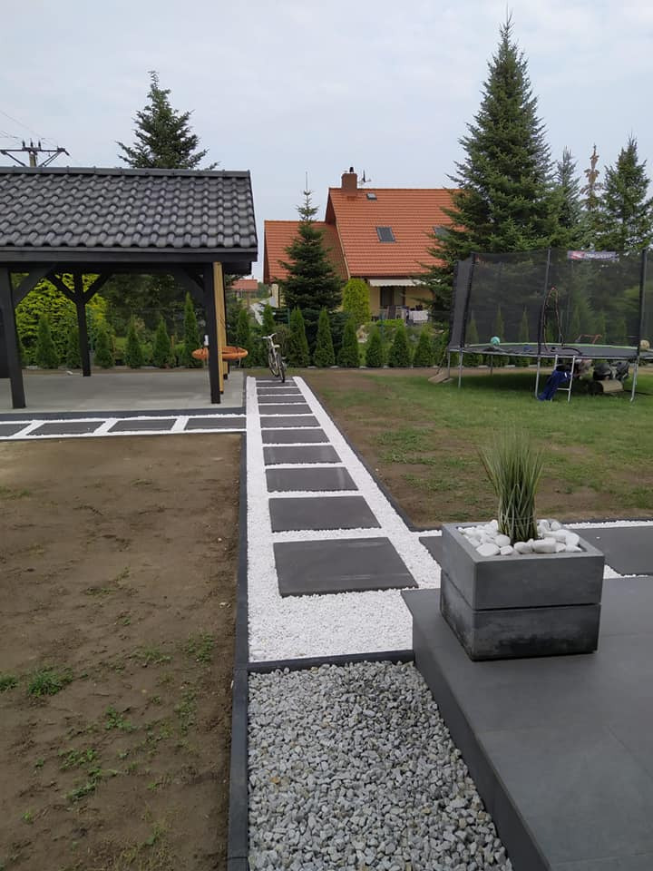 Nowoczesny ogród z ścieżką z ciemnych płyt oddzielonych białym żwirem, donica z ozdobną trawą, altana w tle i trawnik z trampoliną.
