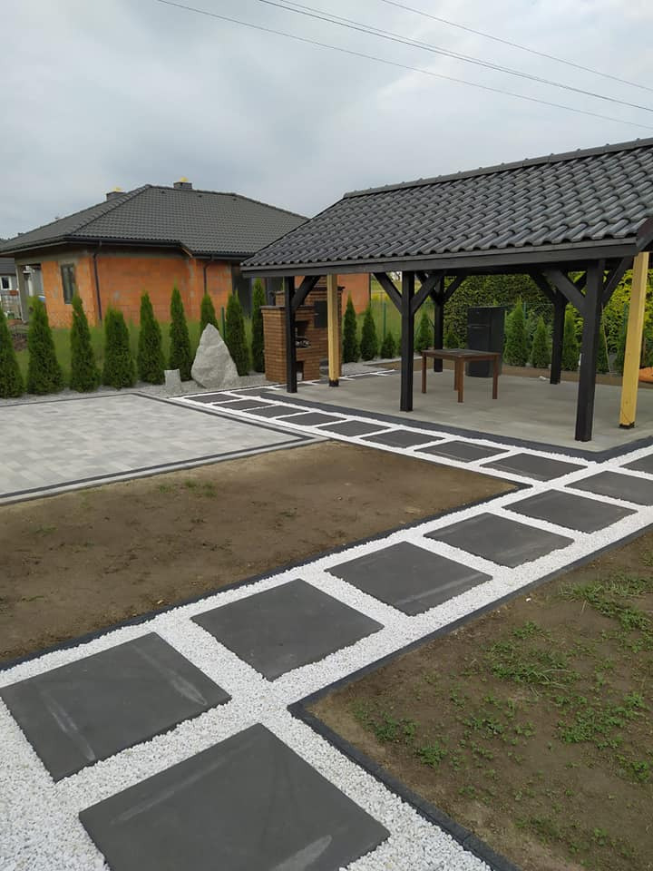 Ogród z nowo ułożoną ścieżką z ciemnych płyt otoczonych białym grysem, prowadzącą do drewnianej altany z grillem murowanym z cegły, widoczny trawnik i budynek w stanie surowym otoczony tujami.