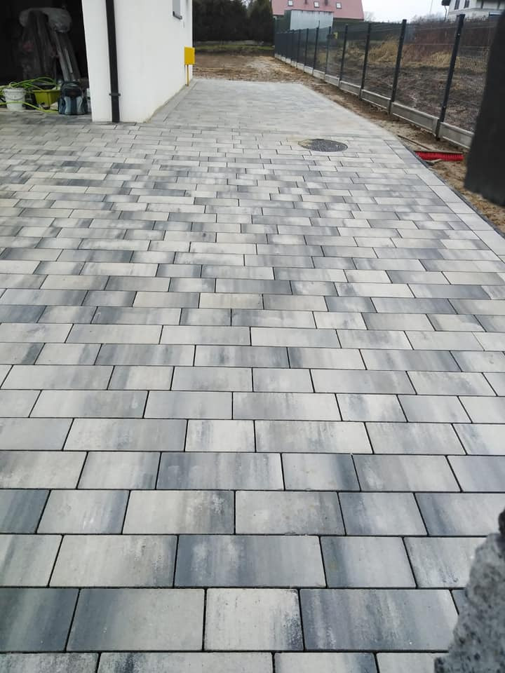 Nowo ułożona, szara kostka brukowa w kształcie prostokątów, prowadząca do białego budynku z żółtą skrzynką gazową, widoczna studzienka kanalizacyjna i ogrodzenie z siatki w tle.