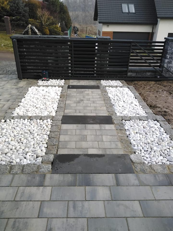 Ułożona ścieżka z szarych kostek brukowych, z obrzeżami z jasnych kamieni i białych otoczaków, prowadząca do czarnej bramy wjazdowej z automatyką.