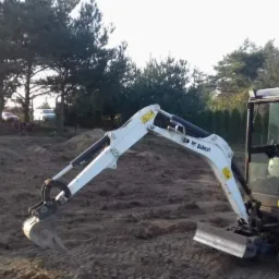 Mini koparka Bobcat E19 na placu budowy, przygotowany teren pod fundamenty, widoczne drzewa w tle.