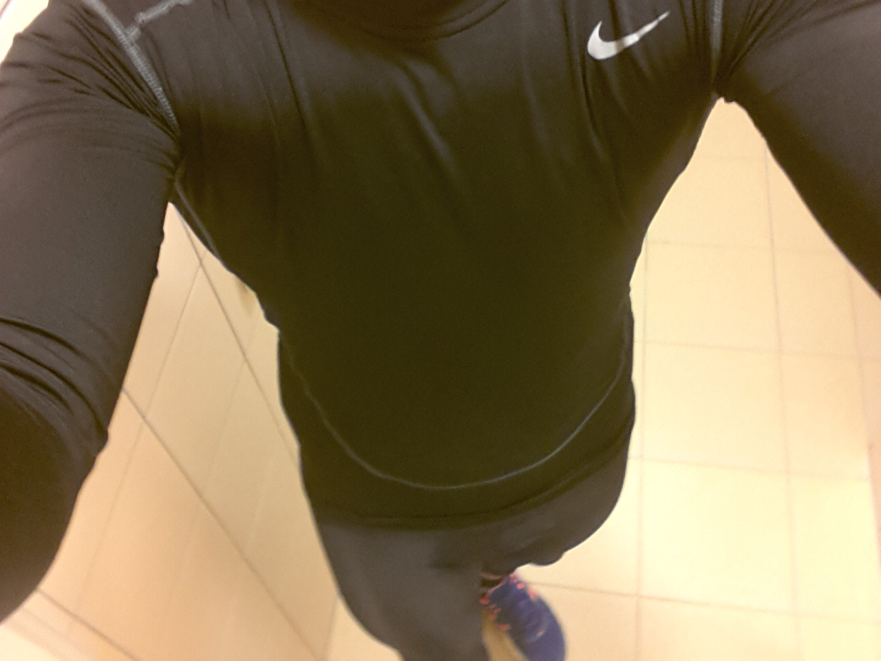 Osoba w czarnej koszulce termoaktywnej z logo Nike i legginsach, widok z góry na kafelkową podłogę, sportowe buty w tle.