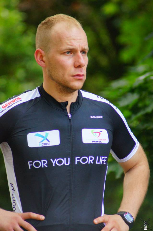 Mężczyzna w sportowej koszulce z logo sponsorów, na tle zieleni, z lekkim zarostem i niebieskimi oczami, patrzy w dal.