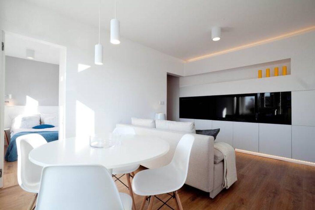 Jasne, minimalistyczne wnętrze mieszkania z okrągłym białym stołem, krzesłami Eames, beżową sofą i zabudową meblową z czarnym panelem na ścianie, widok na sypialnię w tle.