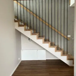 Drewniane schody z czarnymi, metalowymi balustradami i dębowym wykończeniem, na tle szarej ściany z efektem dekoracyjnym. Nowoczesny design.