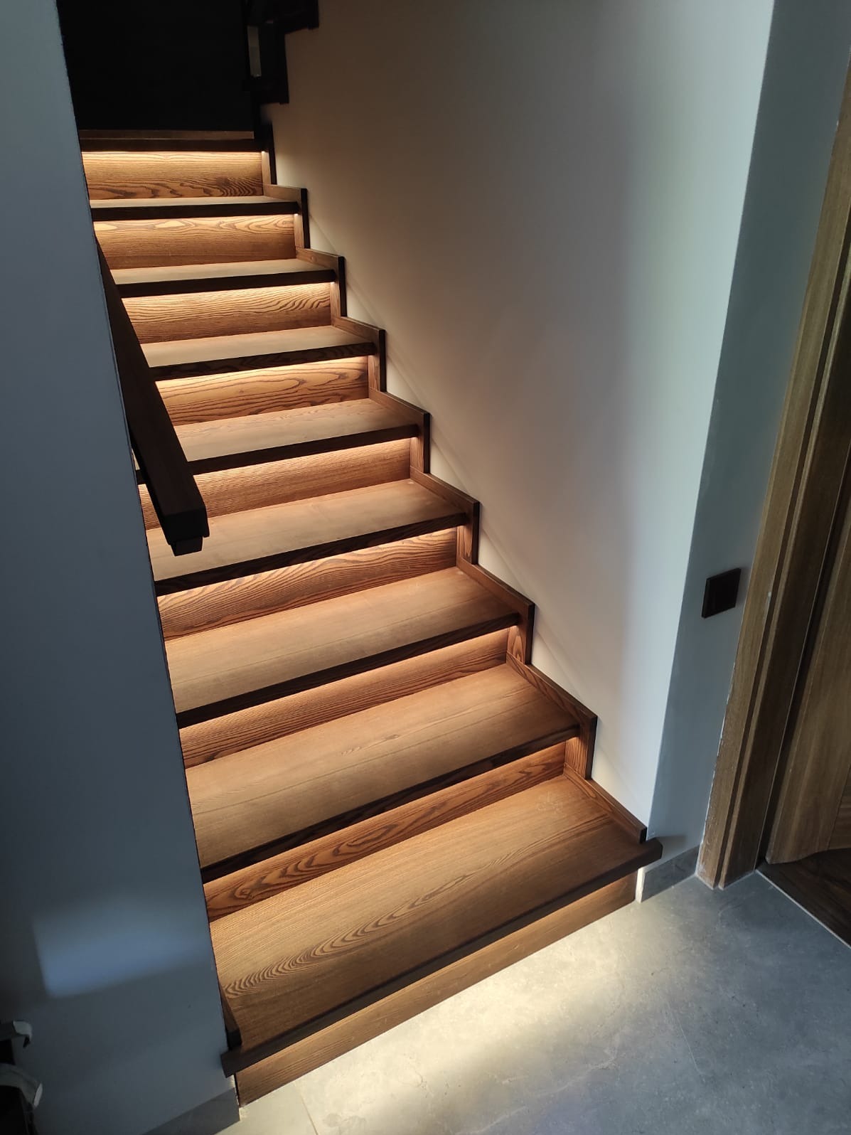 Oświetlone schody z drewna w domu w Wirach. Stopnie podświetlone od spodu, ciemna poręcz po lewej stronie, drzwi po prawej. Widok z dołu na górę.