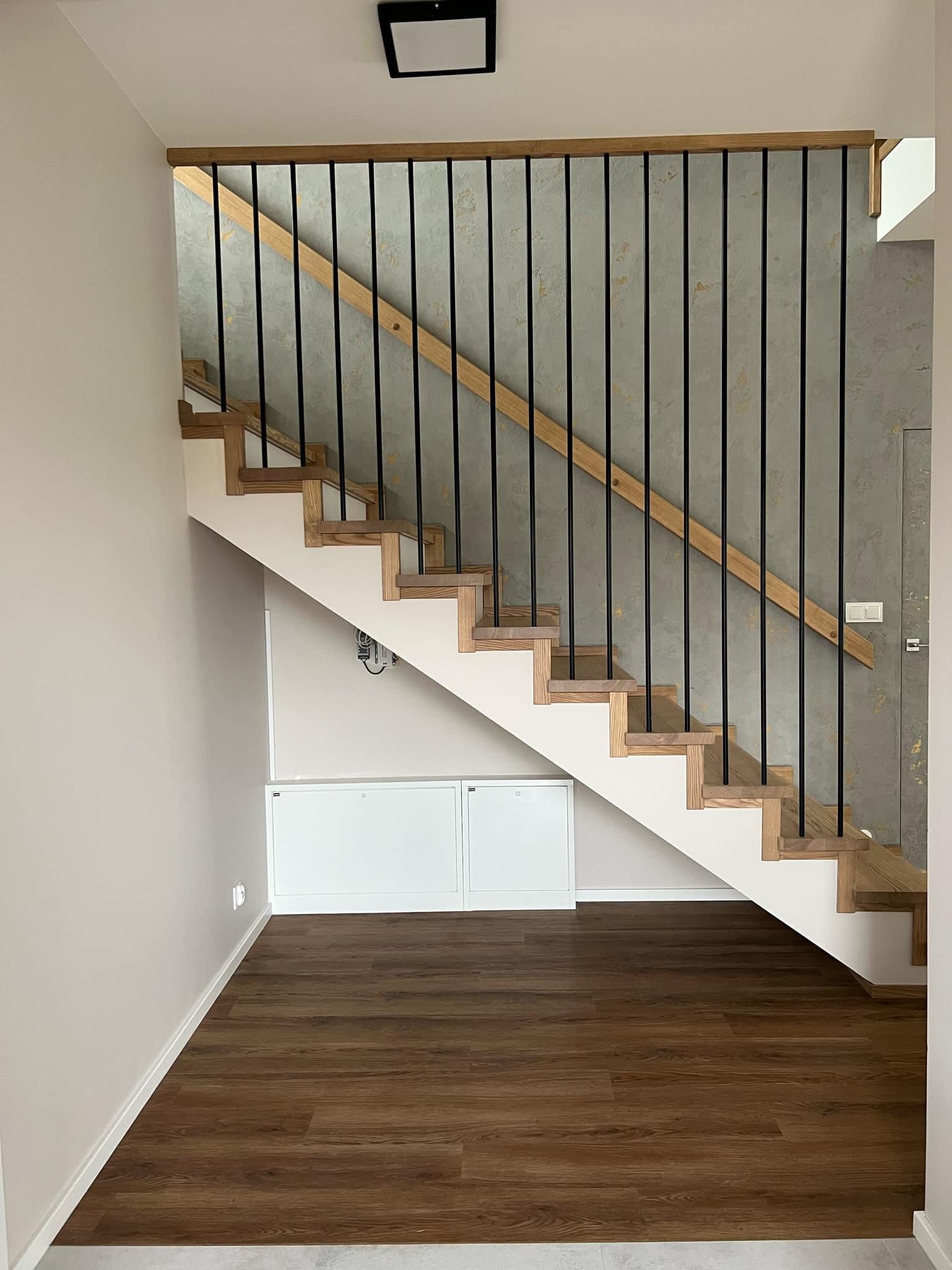 Drewniane schody z czarnymi, metalowymi balustradami i dębowym wykończeniem, na tle szarej ściany z efektem dekoracyjnym. Nowoczesny design.