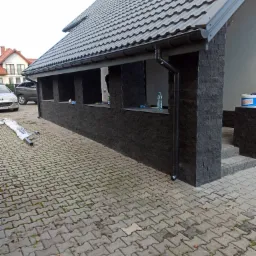 Wykończona elewacja budynku z czarnego kamienia z otworami okiennymi, widoczny fragment dachu z szarą dachówką, rynna spustowa, kostka brukowa na podjeździe i częściowo rozpakowane rury spustowe.