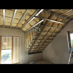 Poddasze w trakcie adaptacji: widoczna konstrukcja metalowa sufitu z izolacją z wełny mineralnej, surowe ściany z oknami, drabina oparta o ścianę.