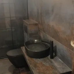 Nowoczesna łazienka z umywalką nablatową w kształcie misy, czarną armaturą i toaletą w kolorze antracytu, wykończona płytkami imitującymi rdzę, z widocznymi przewodami elektrycznymi na ścianie.