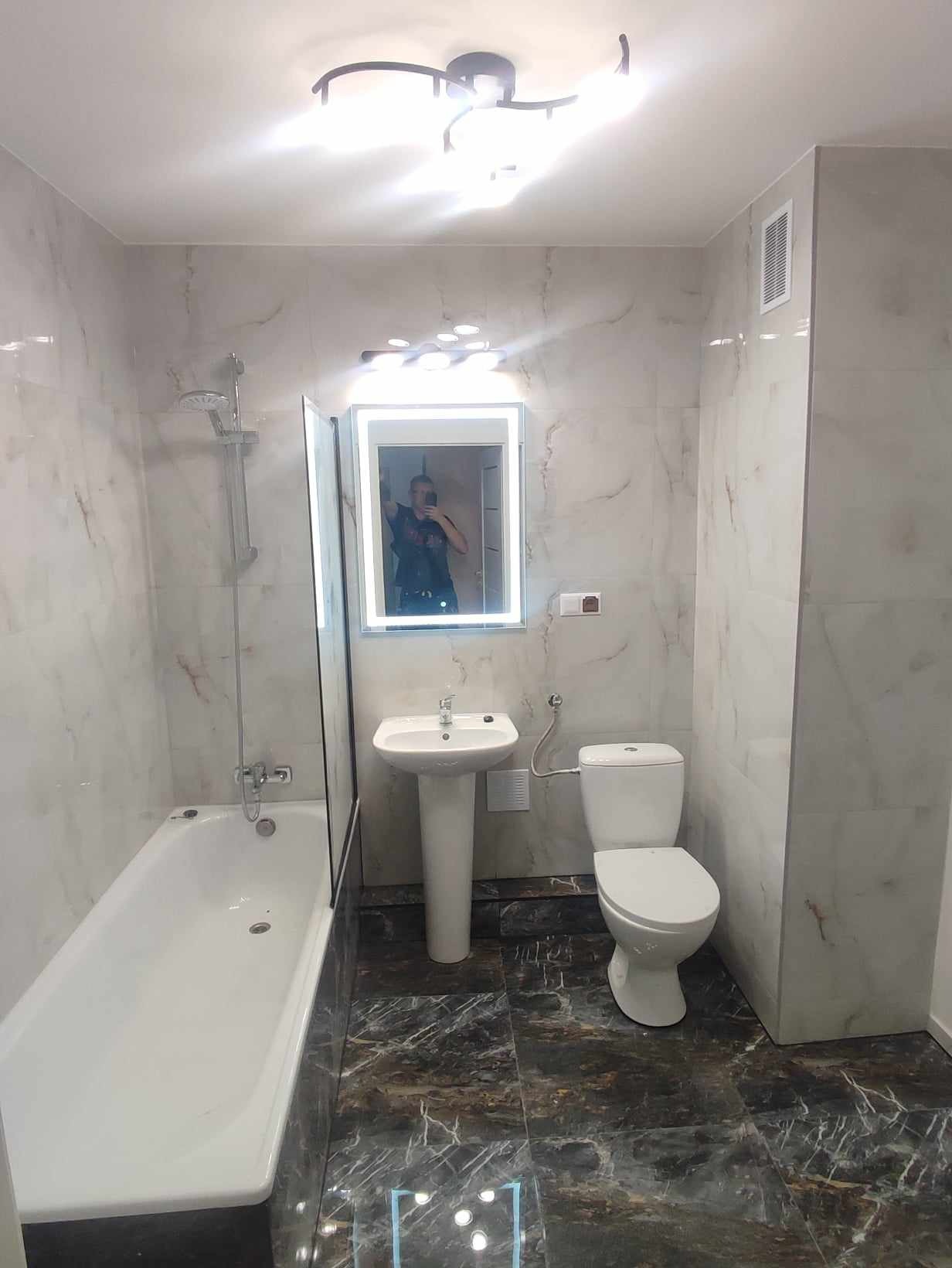 Wykończona łazienka z marmurowymi płytkami na ścianach i podłodze, biała armatura, podświetlane lustro i nowoczesna lampa sufitowa.