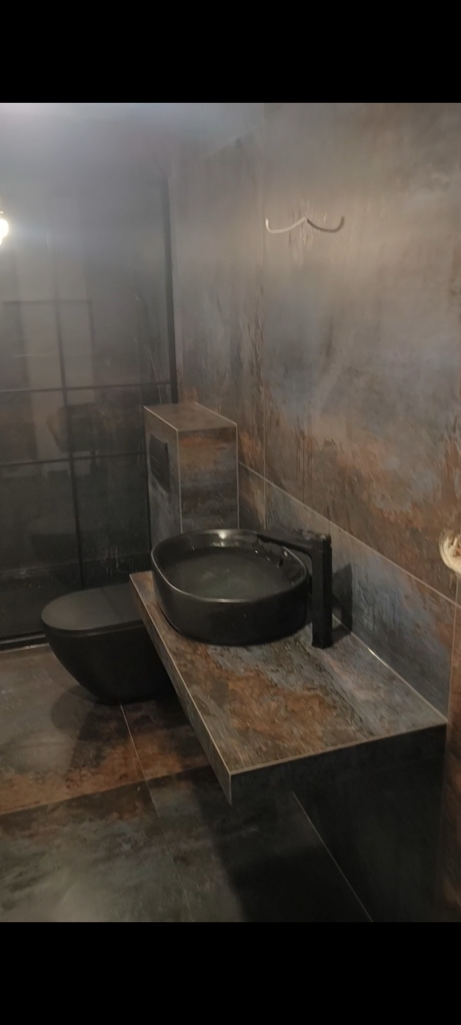 Nowoczesna łazienka z umywalką nablatową w kształcie misy, czarną armaturą i toaletą w kolorze antracytu, wykończona płytkami imitującymi rdzę, z widocznymi przewodami elektrycznymi na ścianie.