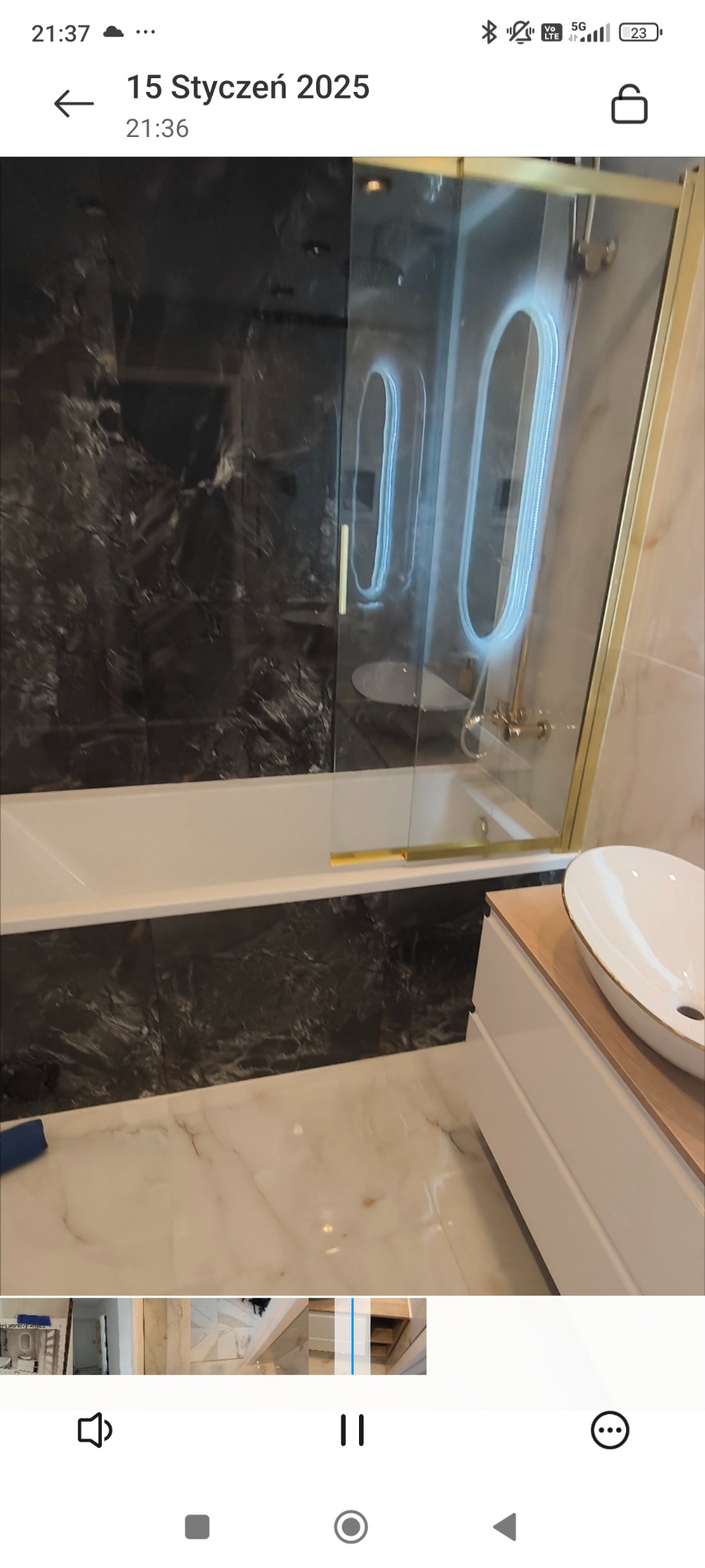 Nowoczesna łazienka z czarnymi marmurowymi płytkami na ścianach, białą wanną, szklanym panelem prysznicowym z złotymi ramami i umywalką nablatową w kształcie elipsy.