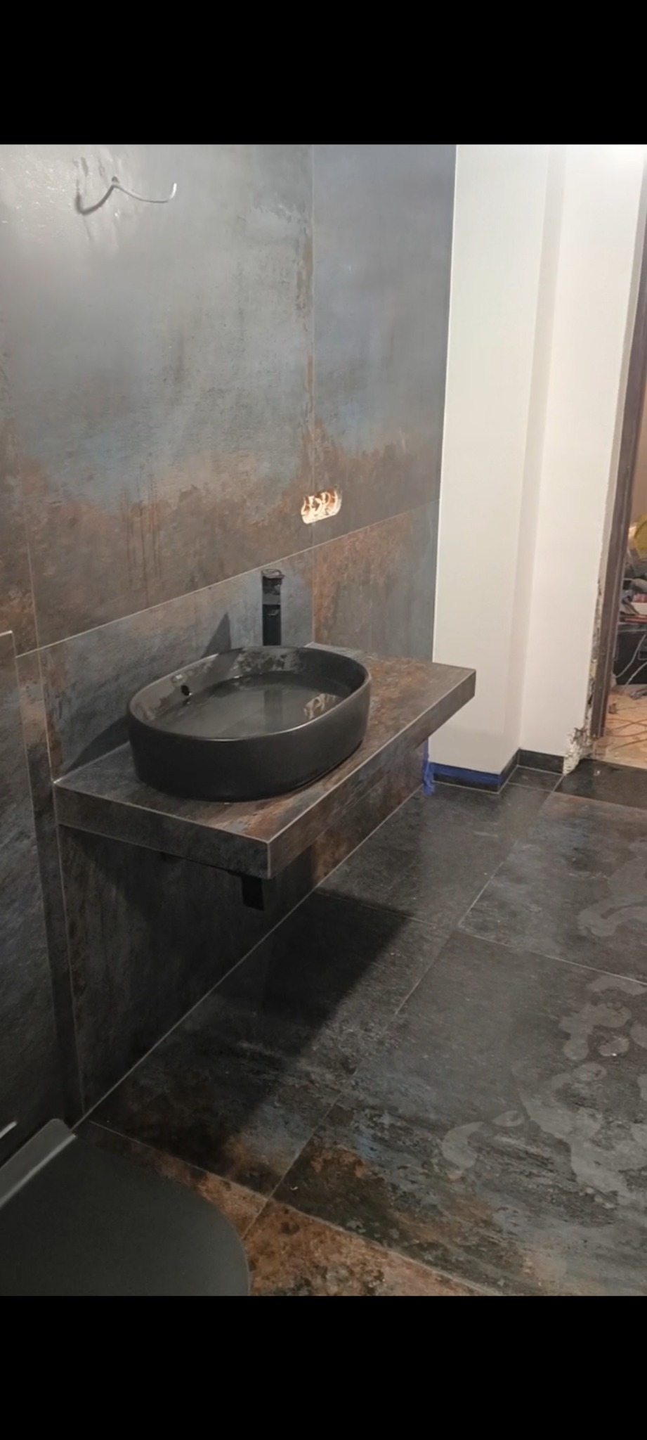 Nowoczesna łazienka w trakcie wykończenia: ciemne płytki imitujące rdzę na ścianach i podłodze, owalna czarna umywalka nablatowa z czarną baterią, widoczne niedokończone prace przy instalacji...