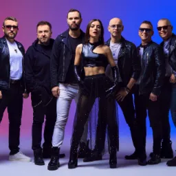 Siedmioosobowa grupa wokalno-instrumentalna pozuje na tle gradientu różu i błękitu, prezentując różnorodne stylizacje, od skórzanych kurtek po prześwitującą spódnicę w gwiazdy i asymetryczny top.