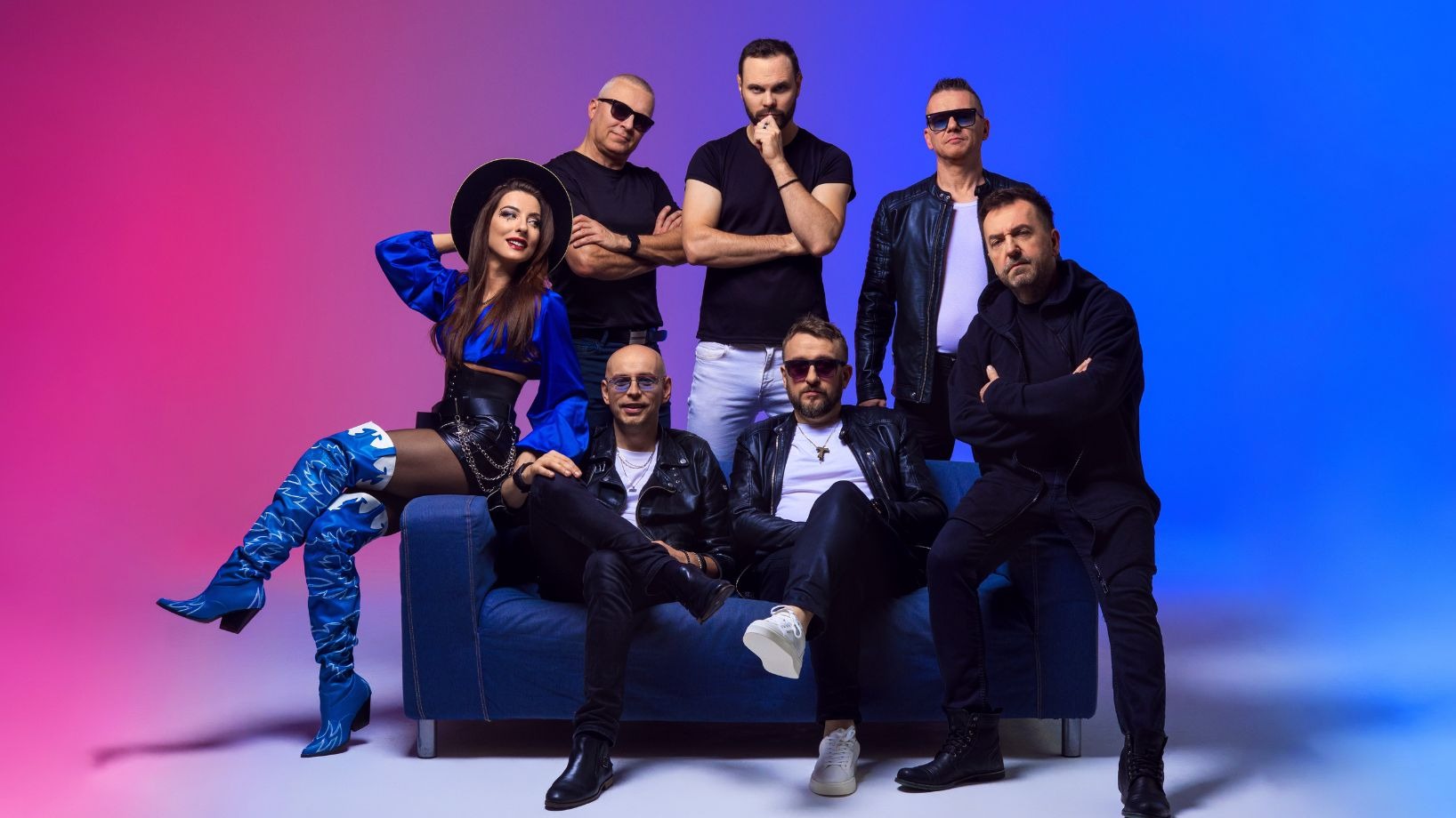 Sześcioosobowa grupa muzyczna w stylizowanych strojach pozuje na niebieskiej kanapie na tle gradientu kolorów, z wokalistką w niebieskich kozakach na pierwszym planie.