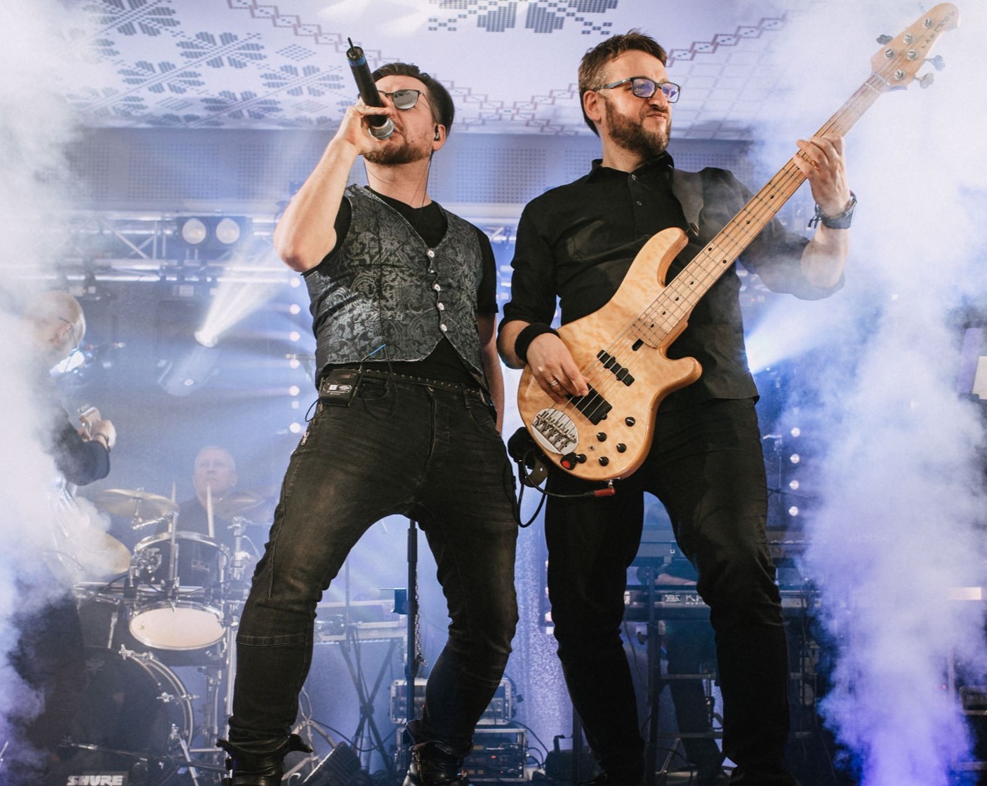 Wokalista w zdobionej kamizelce i basista z jasnym basem na scenie w oparach dymu, perkusja w tle.