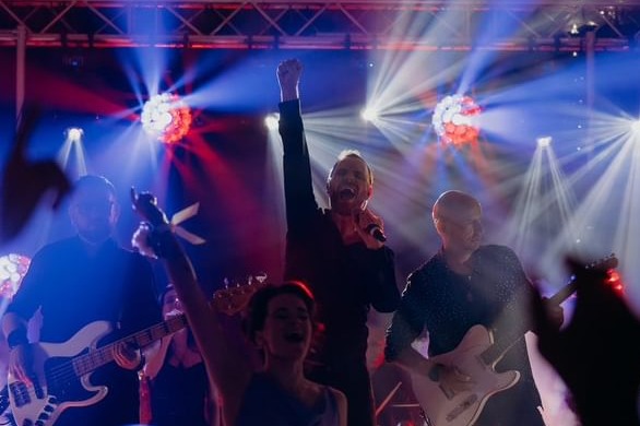 Zespół muzyczny na scenie w trakcie występu, wokalista z podniesioną ręką, oświetlenie sceniczne z kulami dyskotekowymi i reflektorami.