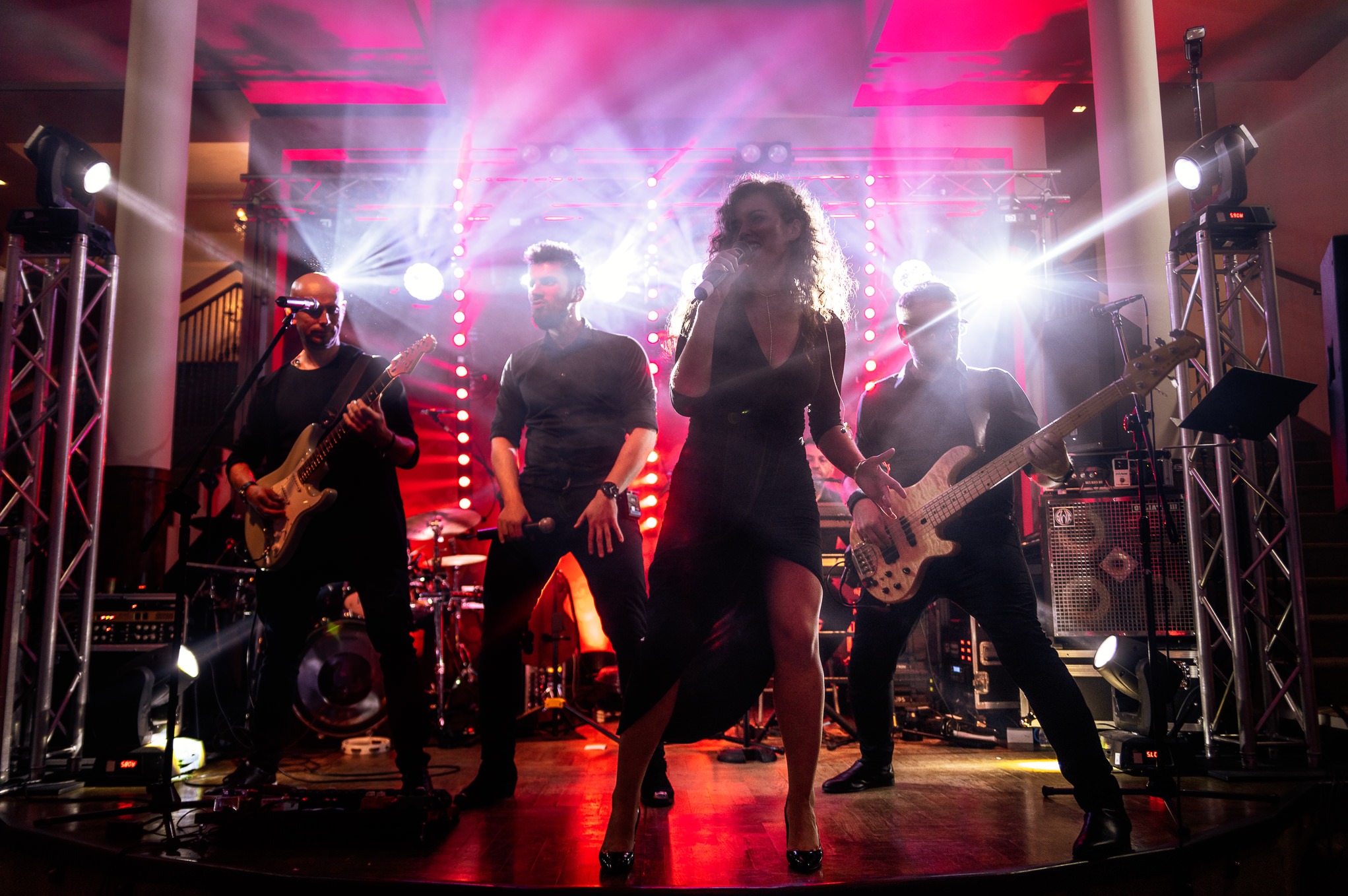 Wokalistka w czarnej sukience śpiewa na scenie z gitarzystami i perkusistą, oświetlona reflektorami i światłami sceny.