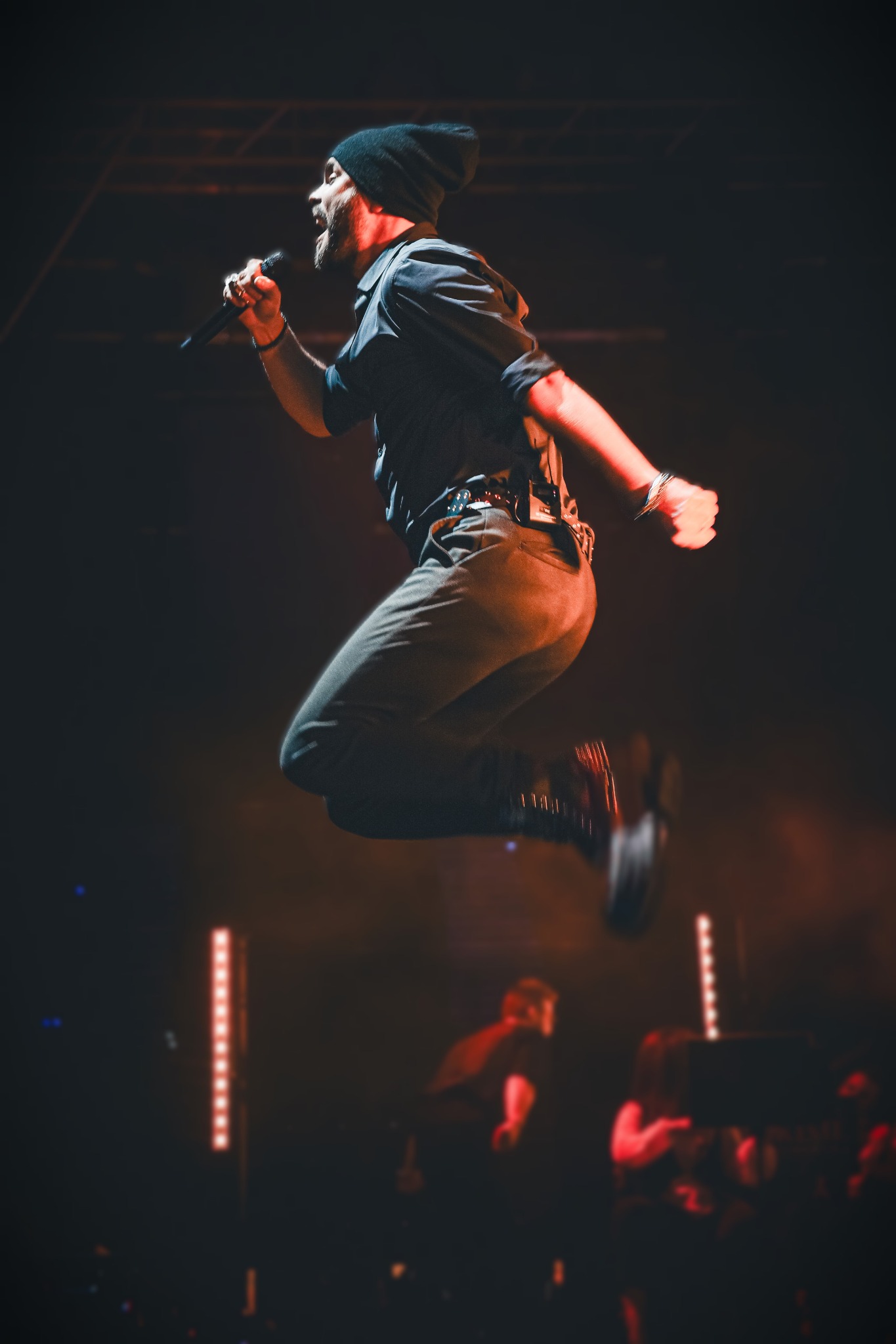 Wokalista w czapce, w skoku, z mikrofonem na scenie podczas występu; czerwone światła w tle.
