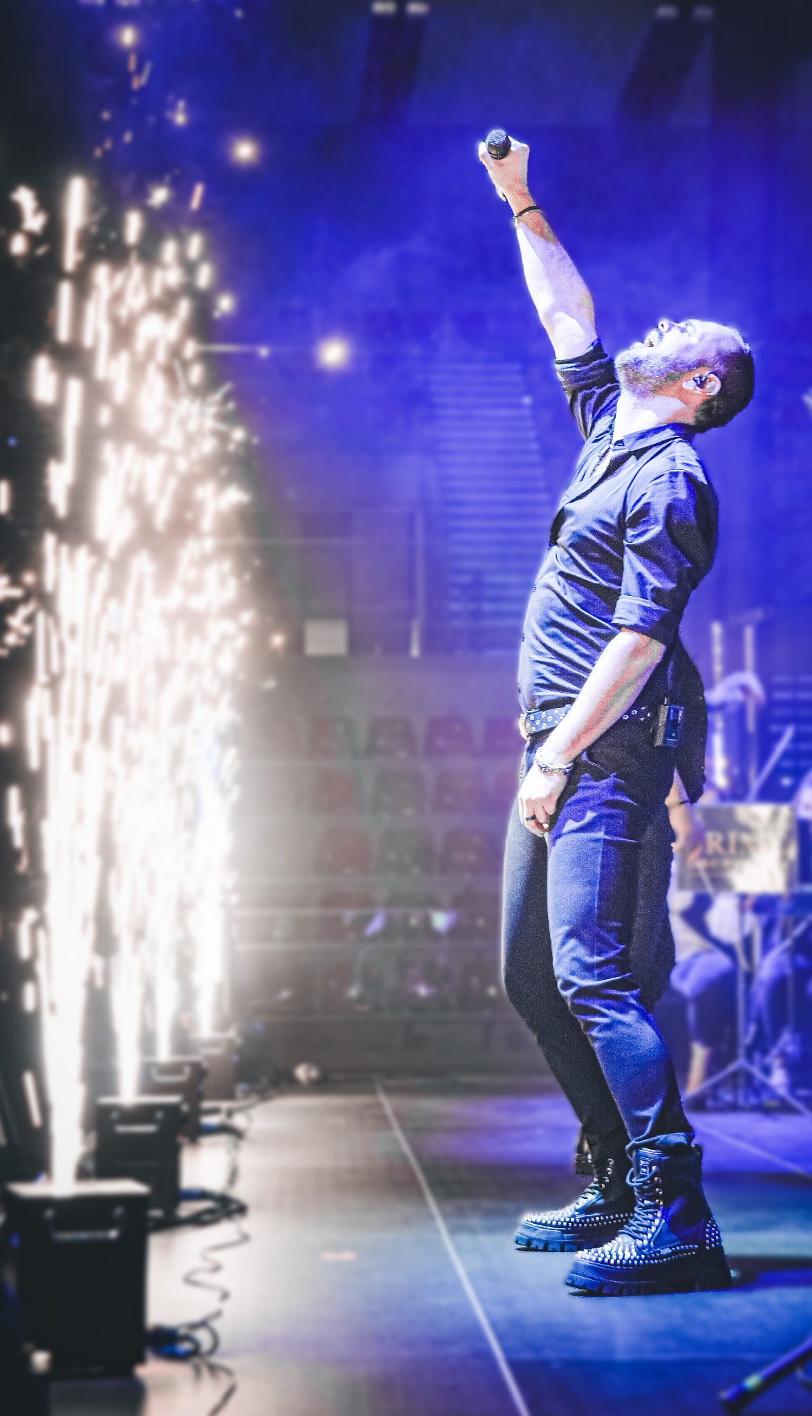 Wokalista z podniesioną ręką trzyma mikrofon na scenie z efektami pirotechnicznymi, widoczne ciężkie, zdobione buty i publiczność w tle.