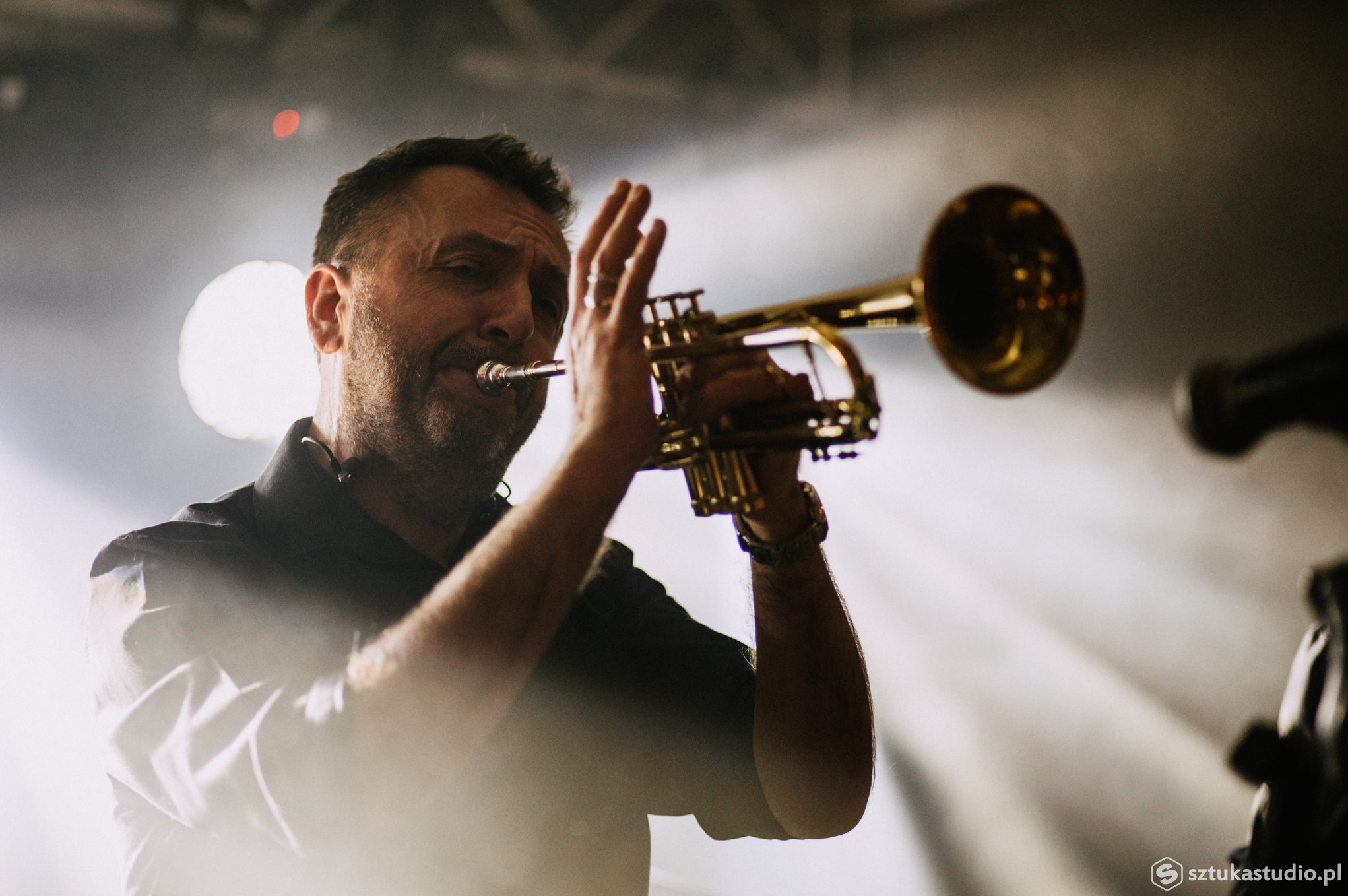 Muzyk grający na trąbce podczas występu scenicznego, w tle rozproszone światło rampy.
